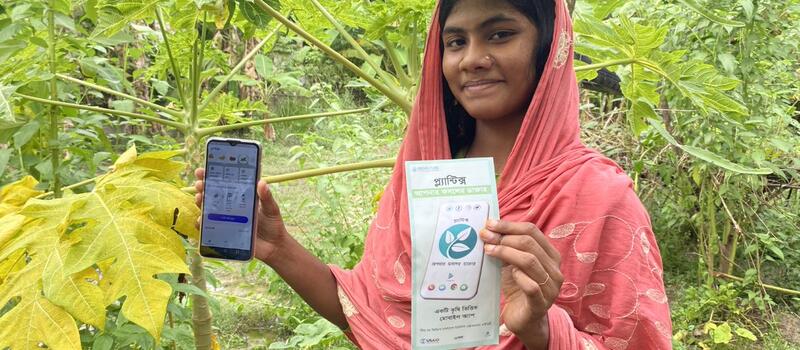 A young woman standing in her farmland displays the mobile application Plantix — Your Crop Doctor